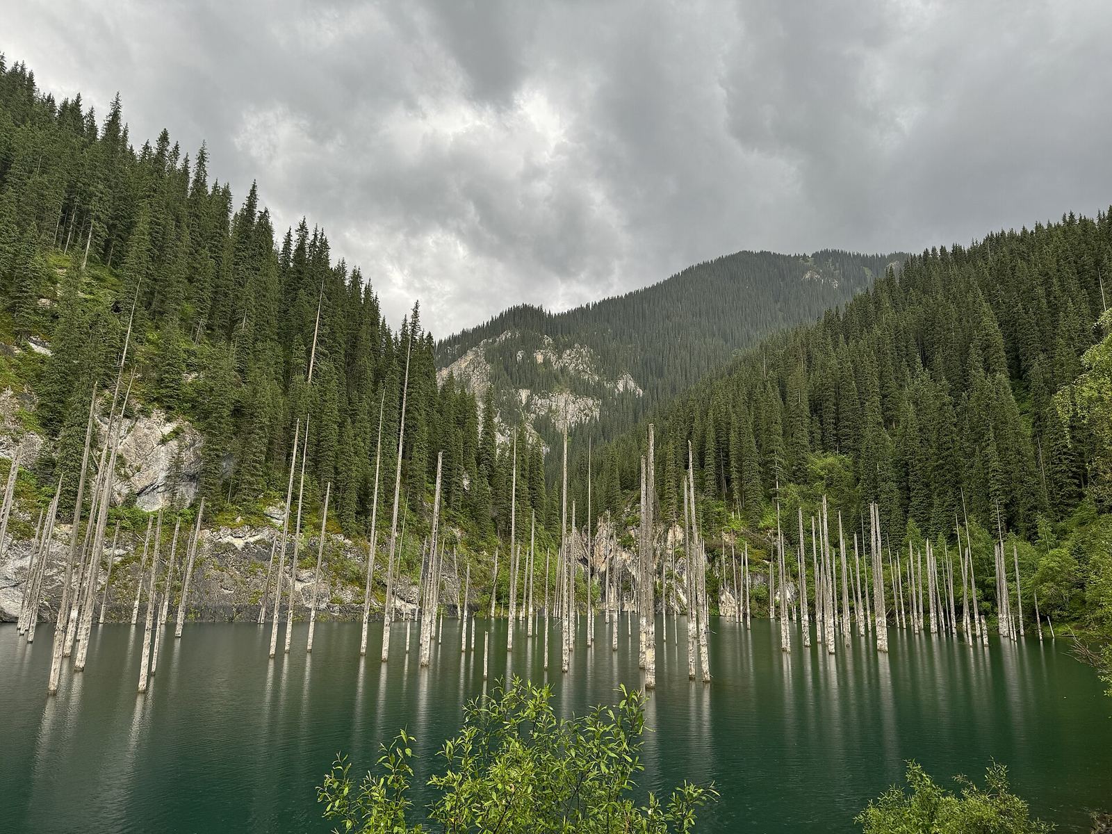 Lake Kaindy underwater forest under a cloudy sky, with spruce-covered slopes behind